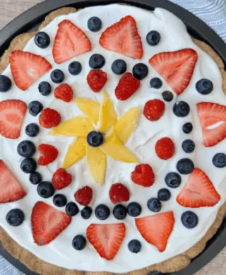 A dessert pizza topped with white frosting and arranged with a starburst of yellow mango slices in the center, surrounded by blueberries, raspberries, and sliced strawberries.