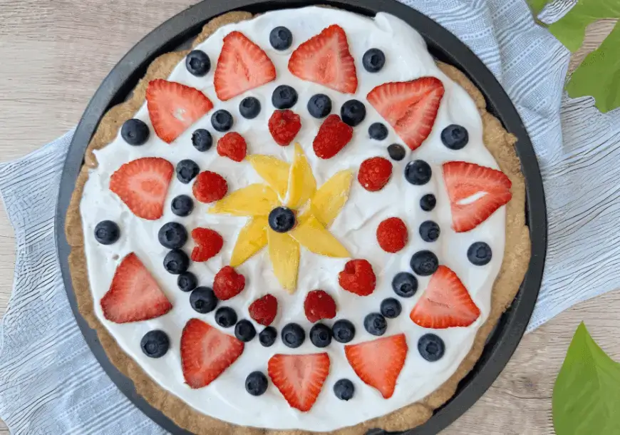 A dessert pizza topped with white frosting and arranged with a starburst of yellow mango slices in the center, surrounded by blueberries, raspberries, and sliced strawberries.