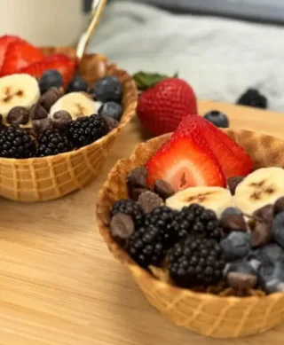 Two waffle bowls filled with yogurt, fruit, and chocolate chips sit on a wooden cutting board.