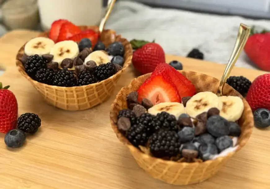 Two waffle bowls filled with yogurt, fruit, and chocolate chips sit on a wooden cutting board.