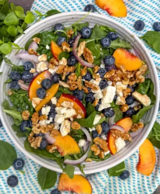 A top-down view of a salad with spinach, blueberries, peaches, red onion, walnuts, and feta cheese in a bowl on a blue and white striped towel.