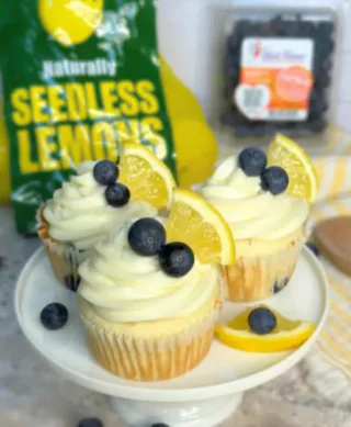 A plate of three cupcakes topped with cream frosting, blueberries, and a slice of lemon, with a bag of seedless lemons in the background.