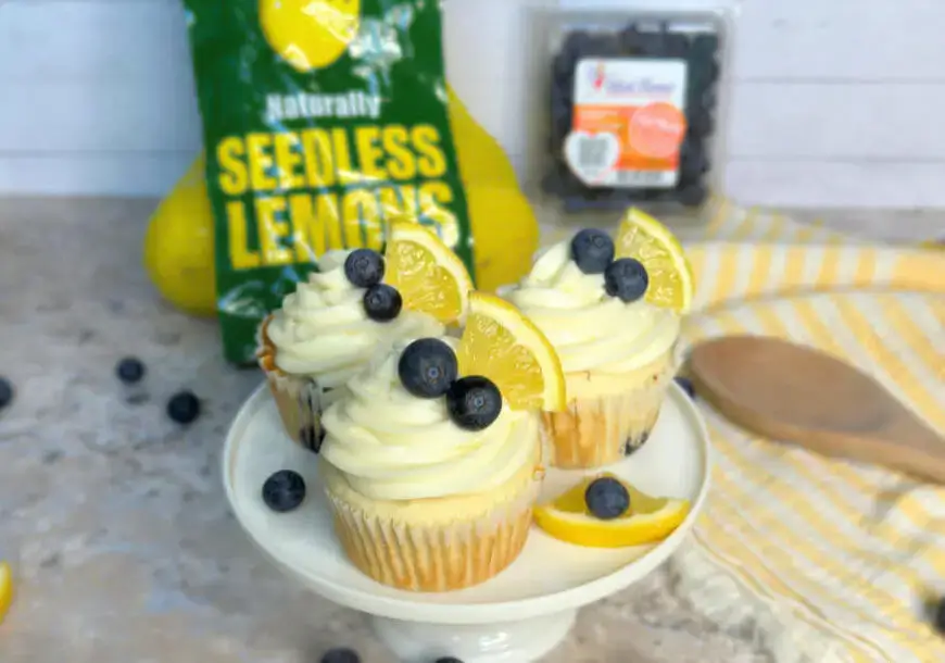 A plate of three cupcakes topped with cream frosting, blueberries, and a slice of lemon, with a bag of seedless lemons in the background.